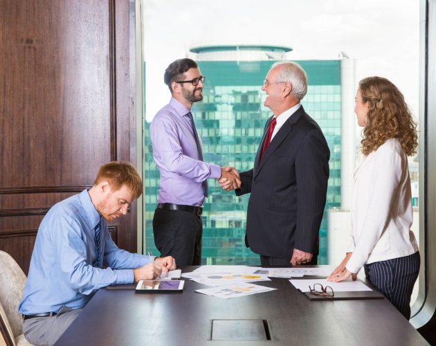 Smiling senior and young businessmen standing at head of table and shaking hands. Businesswoman is standing at table and looking at them. Another businessman is sitting at table and making notes.